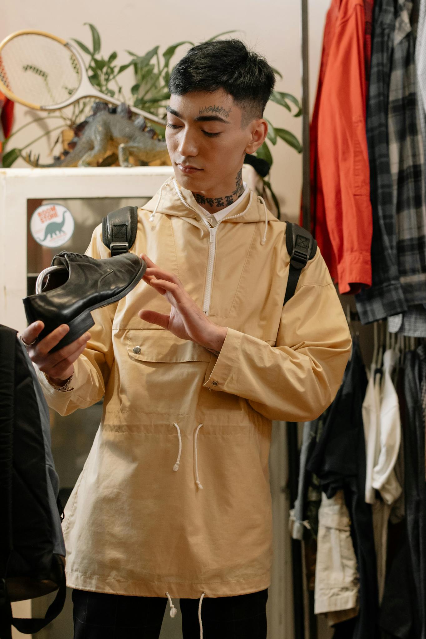 A young man examines a leather shoe in a trendy clothing store, wearing a beige raincoat and backpack.
